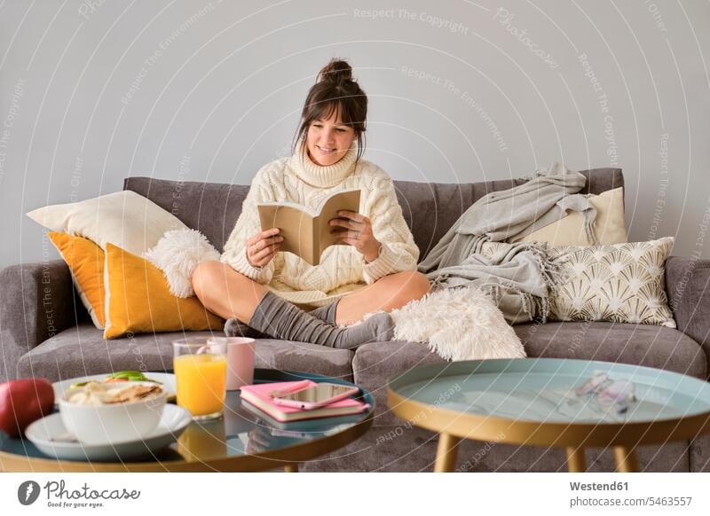 Lächelnde Frau im Pullover lesen Buch beim Sitzen auf dem Sofa zu Hause Farbaufnahme Farbe Farbfoto Farbphoto Spanien Innenaufnahme Innenaufnahmen innen drinnen