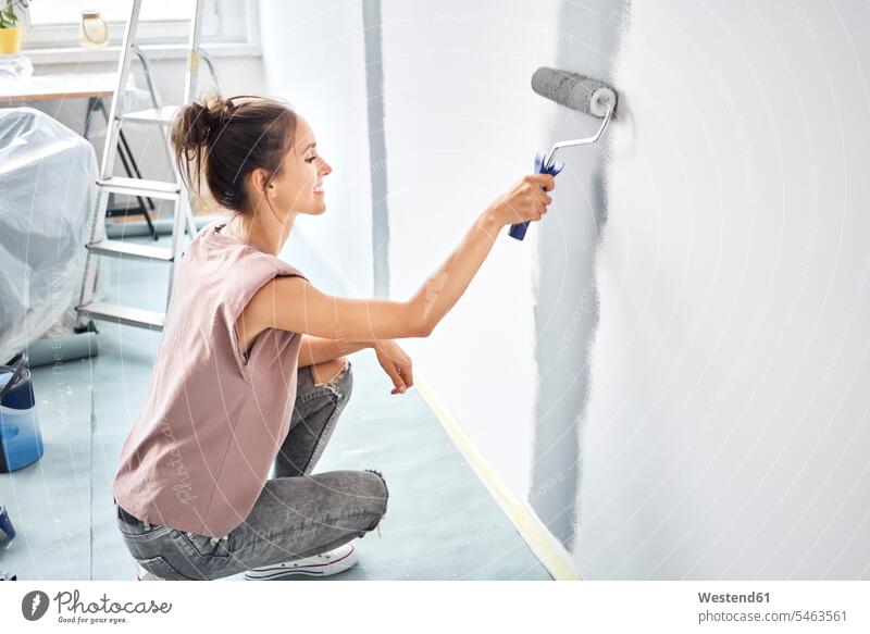 Lächelnde Frau malt mit Farbroller an der Wand, während sie zu Hause hockt Farbaufnahme Farbe Farbfoto Farbphoto Innenaufnahme Innenaufnahmen innen drinnen Tag