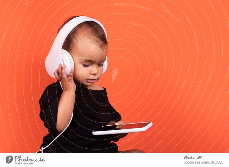 Hübsches Mädchen mit Kopfhörern, das mit seinem Handy telefoniert, während es vor orangem Hintergrund sitzt Farbaufnahme Farbe Farbfoto Farbphoto Studioaufnahme