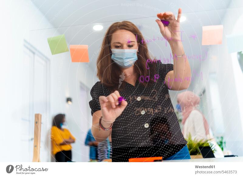 Junge Frau schreibt auf Bildschirmtrennwand, während sie mit einem Kollegen im Hintergrund im Büro steht Farbaufnahme Farbe Farbfoto Farbphoto Innenaufnahme