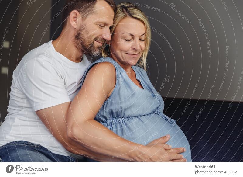 Reifer Mann und seine schwangere reife Frau sitzen auf dem Bett und berühren ihren Bauch Schwangere schwangere Frau sitzend sitzt Betten Bäuche Berührung