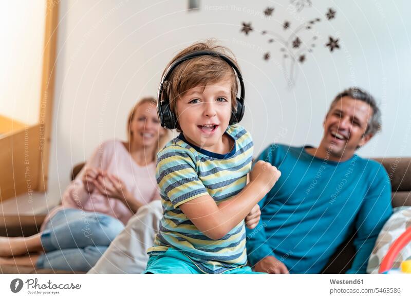 Porträt eines Jungen, der zu Hause auf der Couch mit Kopfhörern Musik hört, mit den Eltern im Hintergrund Couches Liege Sofas Kopfhoerer hoeren begeistert