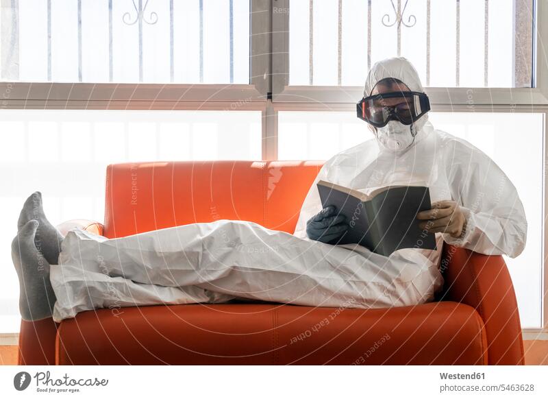 Frau in Schutzkleidung, auf Couch sitzend, Buch lesend Informationen Bücher Masken Couches Liege Sofas Lektüre sitzt weiss weiße weißer weißes stehend steht