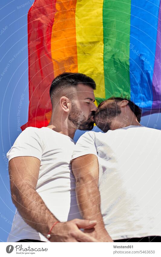 Schwules Paar küsst sich bei Sonnenschein im Stehen unter der Regenbogenflagge Farbaufnahme Farbe Farbfoto Farbphoto Außenaufnahme außen draußen im Freien