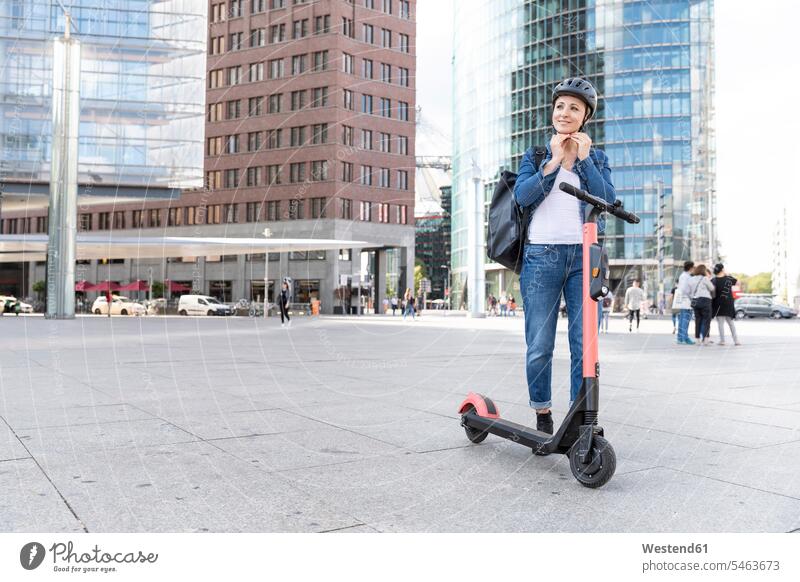 Frau mit E-Scooter in der Stadt, die einen Helm aufsetzt, Berlin, Deutschland verreisen freuen zufrieden stehend steht Baukunst Lifestyles mobil geschützt