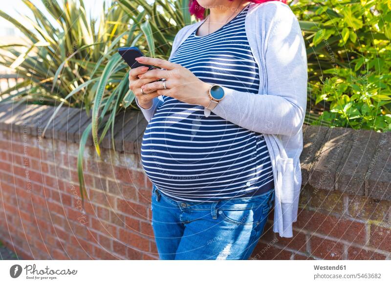 Schwangere Frau tippt draußen auf ihrem Telefon Oberkoerper Oberkörper Torso Torsos Bäuche Telekommunikation telefonieren Handies Handys Mobiltelefon