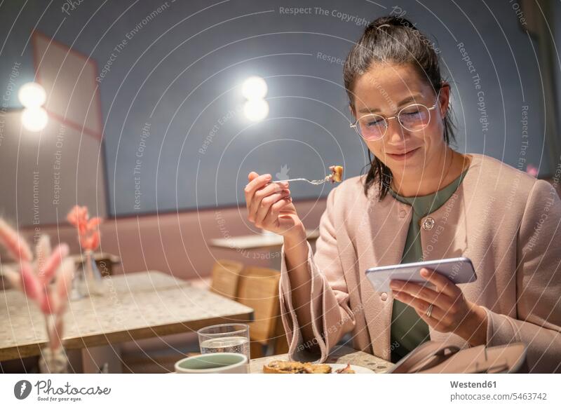 Lächelnde Geschäftsfrau mit Essen, während mit Handy sitzen in modernen Café Farbaufnahme Farbe Farbfoto Farbphoto Innenaufnahme Innenaufnahmen innen drinnen