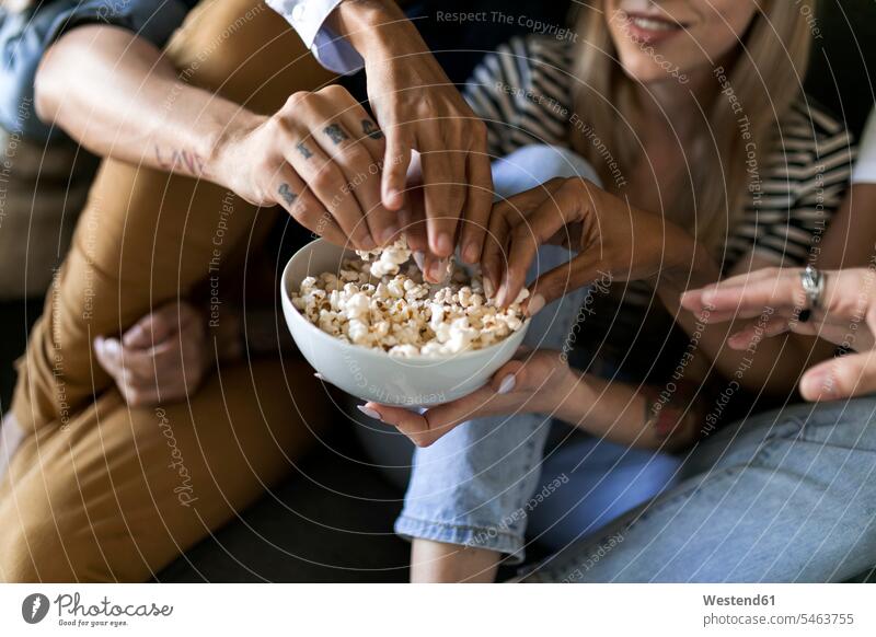 Nahaufnahme von Freunden, die auf der Couch sitzen und sich eine Schüssel Popcorn teilen sitzend sitzt essen essend Puffmais Schalen Schälchen Schüsseln Sofa