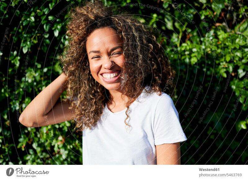 Fröhliche junge Frau, die bei Sonnenschein mit der Hand im Haar gegen Pflanzen steht Farbaufnahme Farbe Farbfoto Farbphoto Außenaufnahme außen draußen im Freien