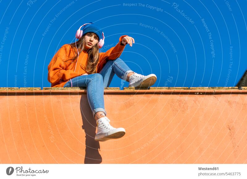 Porträt eines Teenager-Mädchens, das an der Wand vor dem Himmel sitzt und mit Kopfhörern Musik hört Kopfhoerer hoeren sitzend Farben Farbtoene Farbton Farbtöne