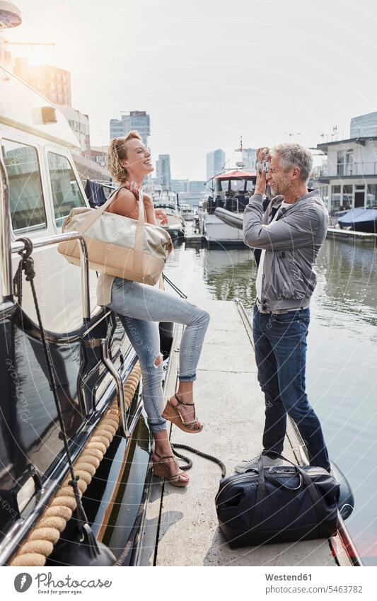 Älterer Mann, der eine junge Frau auf einem Steg neben einer Jacht fotografiert Stege Anlegestelle stehen stehend steht Reisetasche Reisetaschen Yacht Yachten