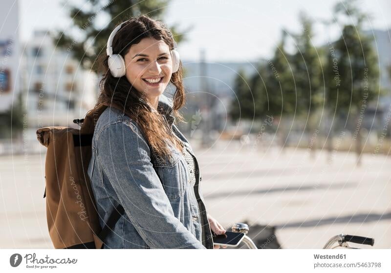 Lächelnde Studentin hört Musik über Kopfhörer und steht an einem sonnigen Tag in der Stadt Farbaufnahme Farbe Farbfoto Farbphoto Deutschland