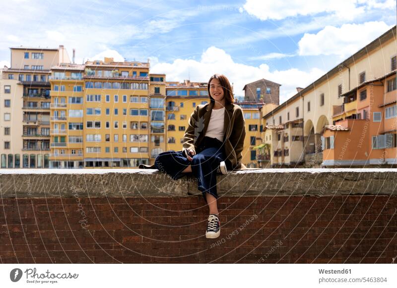 Italien, Florenz, lächelnde junge Frau, die sich auf einer Mauer in der Stadt ausruht Wohnhaus Wohnhäuser Wohnhaeuser wohnen Portrait Porträts Portraits positiv