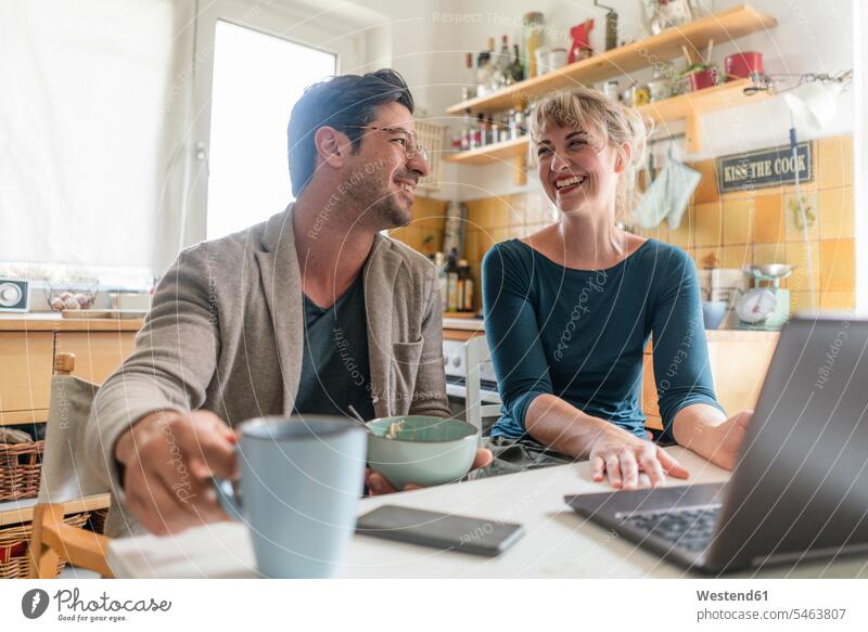 Glückliches Paar sitzt in der Küche mit Laptop am Tisch Schale Schalen Schälchen Schüsseln Rechner Laptops Notebook Notebooks Telekommunikation telefonieren