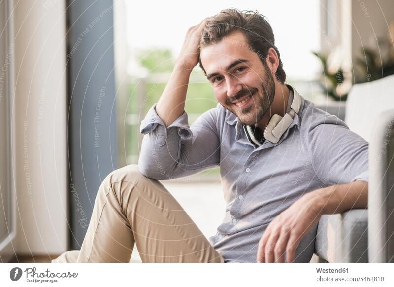 Porträt eines jungen Mannes, der in seinem Wohnzimmer sitzt, mit der Hand im Haar Europäer Kaukasier Europäisch kaukasisch freundlich nett offenes Lächeln