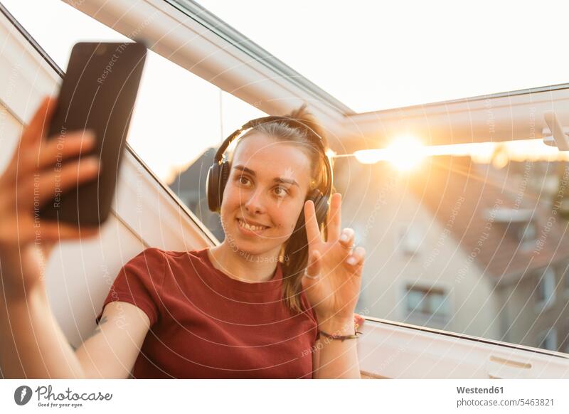 Junge Frau bei einem Videochat am Fenster im Gegenlicht T-Shirts Kopfhoerer Telekommunikation telefonieren Handies Handys Mobiltelefon Mobiltelefone abends
