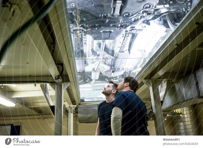 Zwei Kollegen in der Fabrik überprüfen Unterboden eines Autos Arbeitskollege Arbeitskollegen Job Berufe Berufstätigkeit Beschäftigung Jobs Arbeiter T-Shirts KFZ