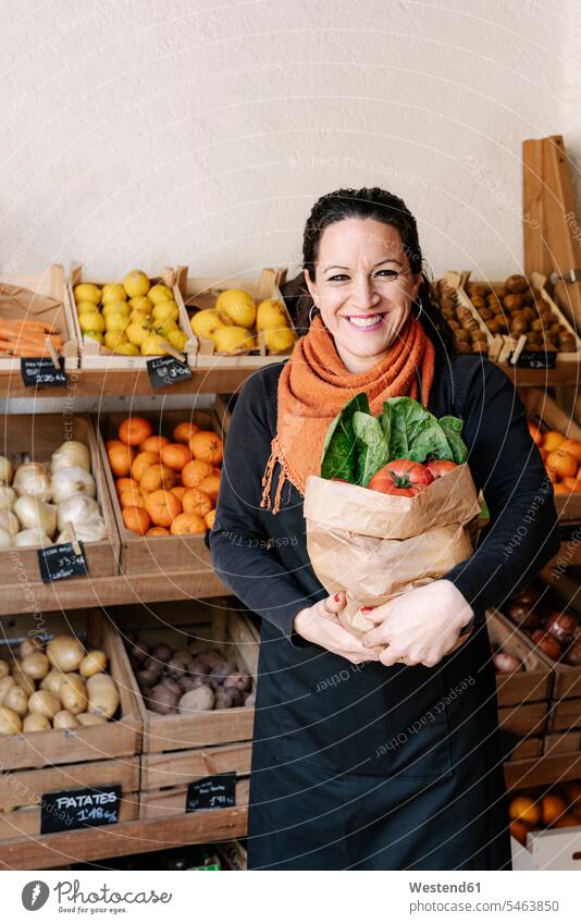 Fröhliche Verkäuferin hält Tüte mit Gemüse und steht im Laden gegen Kartons Frische frisch Gesunde Ernährung gesund Gesundheit Food and Drink Essen und Trinken