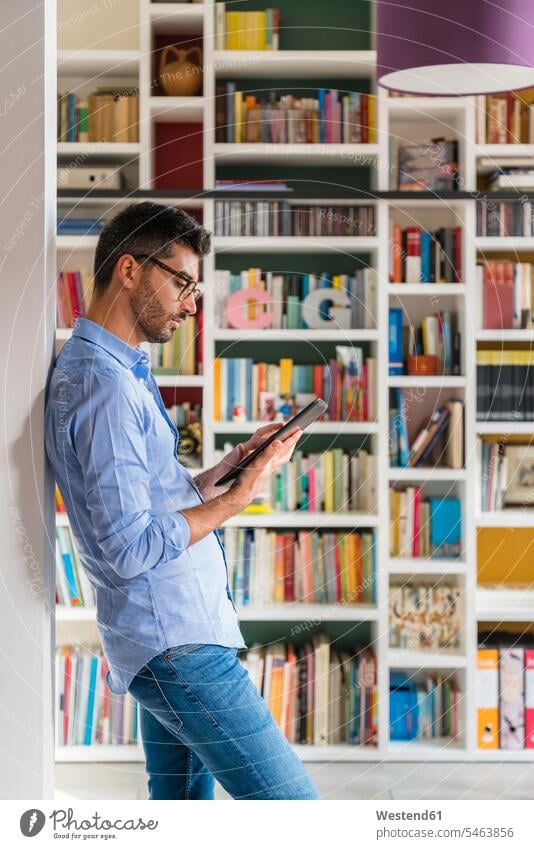Junger Mann steht zu Hause mit digitalem Tablet vor dem Bücherregal Leute Menschen People Person Personen Europäisch Kaukasier kaukasisch 1 Ein ein Mensch eine