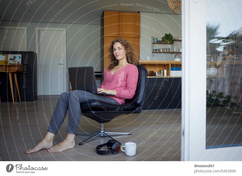 Mittlere erwachsene Frau, die am Laptop arbeitet, während sie zu Hause auf einem Stuhl sitzt Farbaufnahme Farbe Farbfoto Farbphoto Außenaufnahme außen draußen