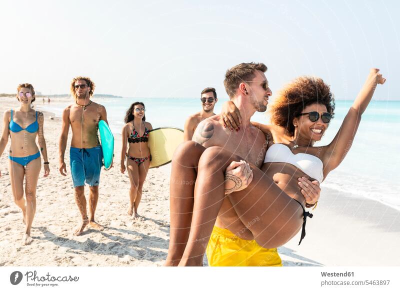 Freunde gehen am Strand spazieren, ein Mann trägt seine Freundin gehend geht Surfbrett Surfbretter surfboard surfboards Surfer Wellenreiter Beach Straende