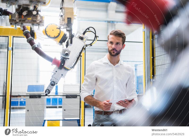 Zuversichtlicher Ingenieur schaut auf Roboterarm, während er mit digitalem Tablett in der Fabrik steht Farbaufnahme Farbe Farbfoto Farbphoto Deutschland