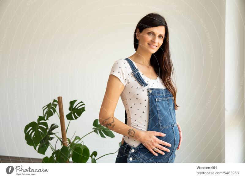 Lächelnde schwangere Frau mit Händen auf dem Bauch an der Wand stehend in neuem Zuhause Farbaufnahme Farbe Farbfoto Farbphoto Deutschland Innenaufnahme