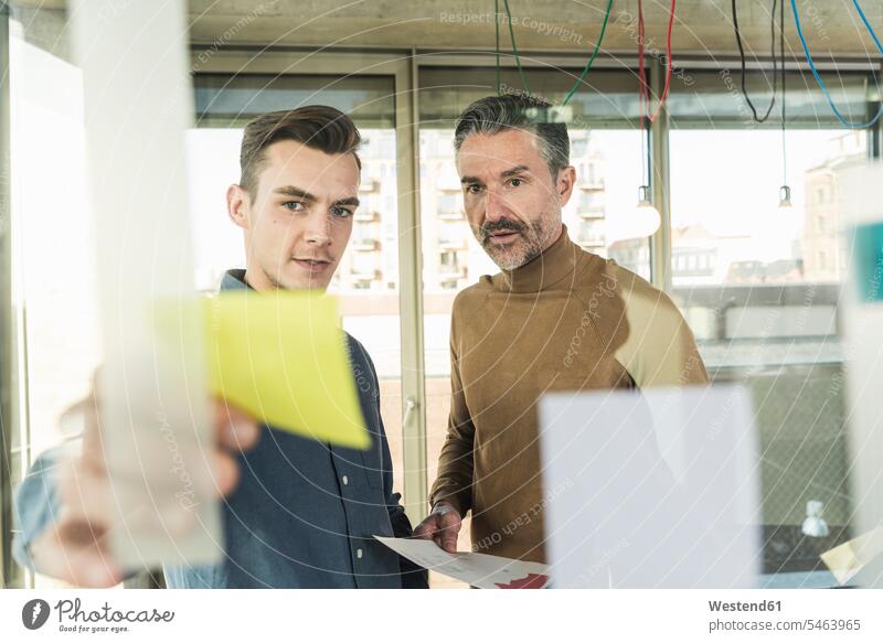 Reifer Geschäftsmann und junger Mann arbeiten an Haftnotizen an einer Glasscheibe im Büro Arbeitskollege Arbeitskollegen Kollege Partner Partnerschaften Job