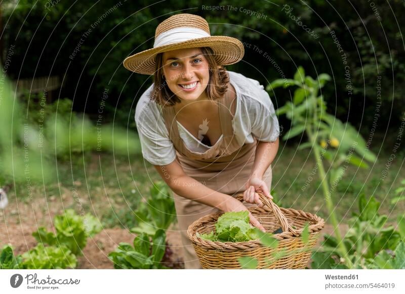 Lächelnde junge Frau mit Hut sammelt Gemüse im Weidenkorb auf dem Hof Farbaufnahme Farbe Farbfoto Farbphoto Spanien Latzhose Außenaufnahme außen draußen