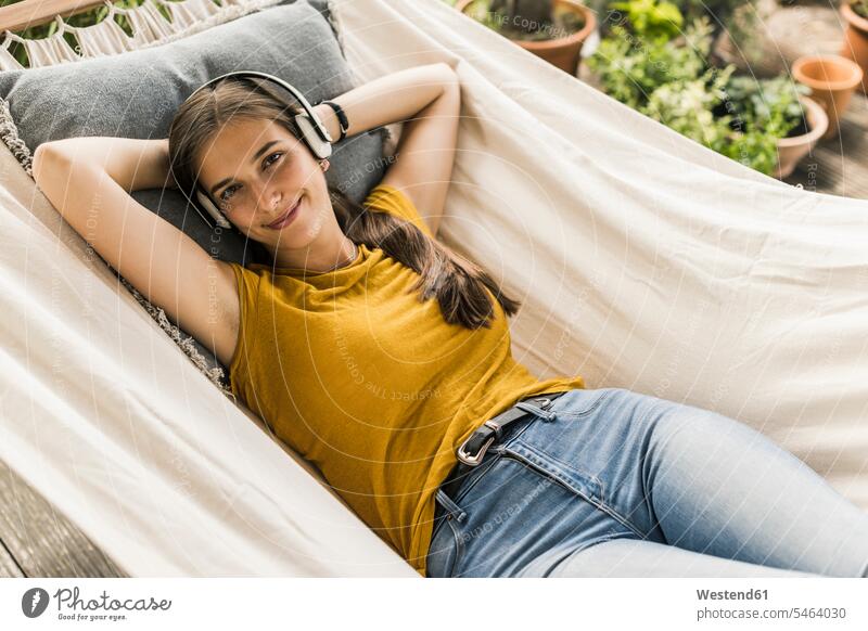 Lächelnde junge Frau hört Musik über Kopfhörer, während sie auf einer Hängematte im Hof liegt Farbaufnahme Farbe Farbfoto Farbphoto Deutschland