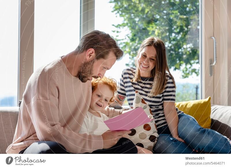 Lächelnde Frau sitzt und schaut den Mann an, der dem Jungen im Wohnzimmer ein Bilderbuch vorliest Farbaufnahme Farbe Farbfoto Farbphoto Innenaufnahme