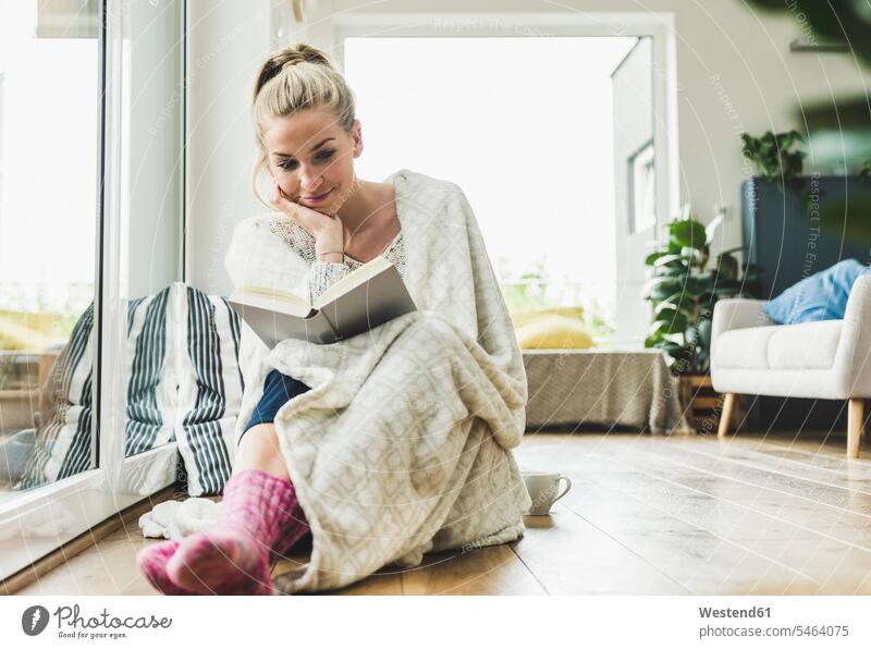 Frau eingewickelt in eine Decke sitzt am Fenster zu Hause ein Buch lesen Leute Menschen People Person Personen Alleinstehende Alleinstehender Singles