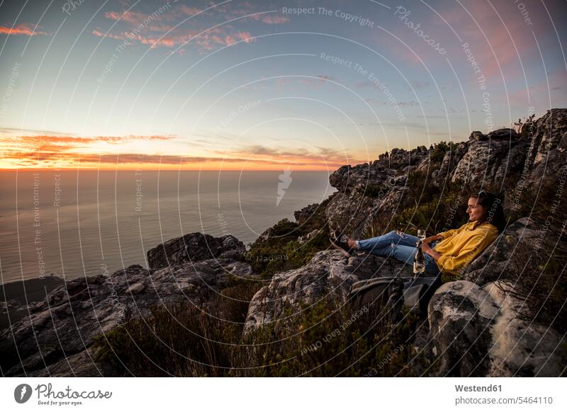 Südafrika, Kapstadt, Tafelberg, Frau sitzt auf einem Felsen und trinkt Wein bei Sonnenuntergang weiblich Frauen Sonnenuntergänge sitzen sitzend trinken Weine