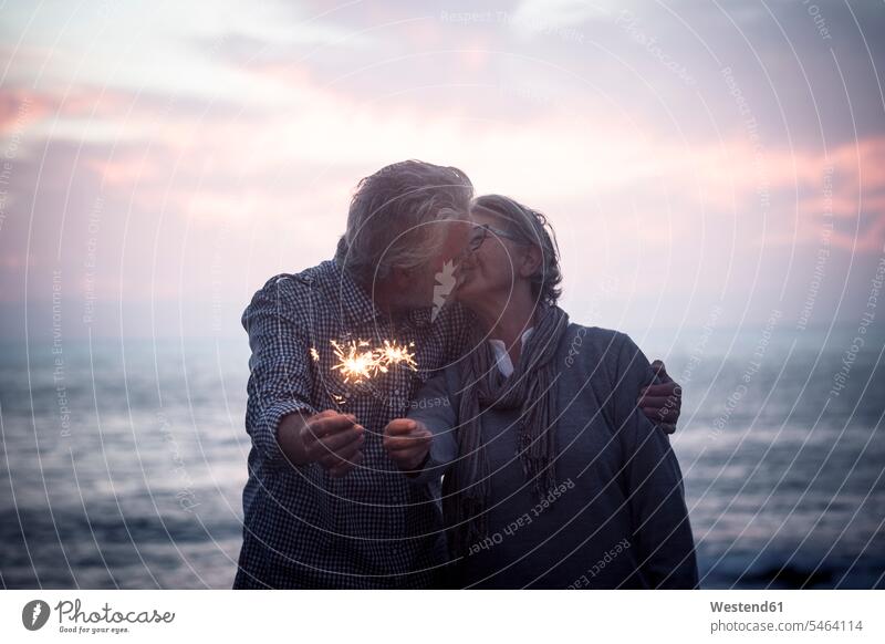 Küssendes älteres Paar steht vor dem Meer bei Sonnenuntergang und hält Wunderkerzen Spanien Abendstimmung Vignettierung Abschattung Stimmungsvoller Himmel