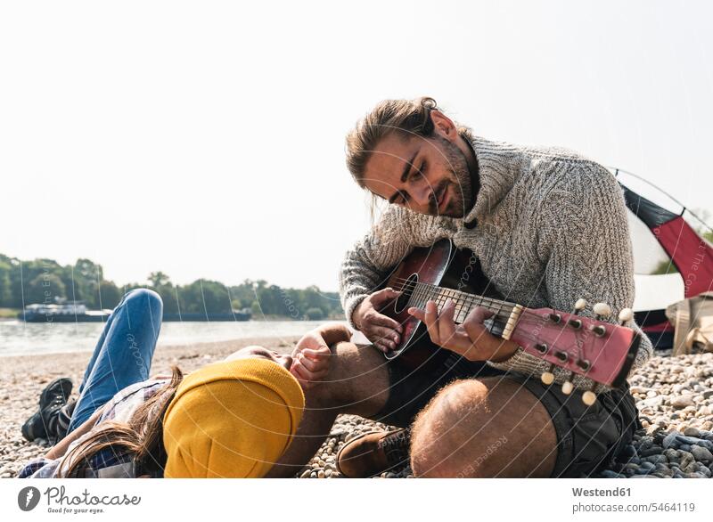 Glückliches junges Paar spielt Gitarre in einem Zelt am Flussufer Gitarren Pärchen Paare Partnerschaft Zelte Saiteninstrument Saiteninstrumente Musikinstrument
