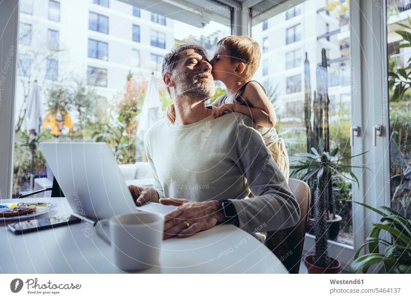Kleiner Junge umarmt Vater von hinten, während er von zu Hause aus arbeitet Job Berufe Berufstätigkeit Beschäftigung Jobs Tische Rechner Laptops Notebook