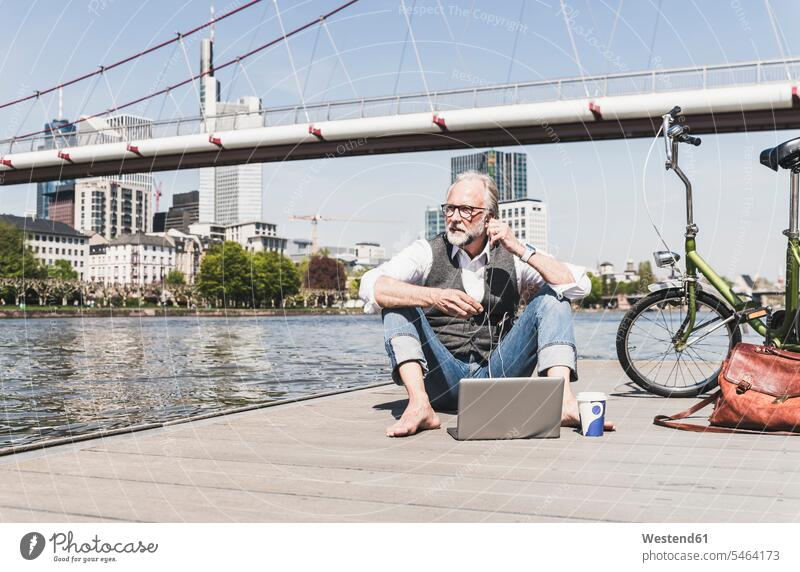 Reifer Mann mit Laptop, Ohrstöpseln und Fahrrad am Flussufer in der Stadt sitzend staedtisch städtisch Bikes Fahrräder Räder Rad sitzt Männer männlich Notebook
