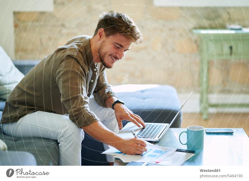 Lächelnder Geschäftsmann schreibt auf Papier, während er im Wohnzimmer am Laptop sitzt Farbaufnahme Farbe Farbfoto Farbphoto Innenaufnahme Innenaufnahmen innen