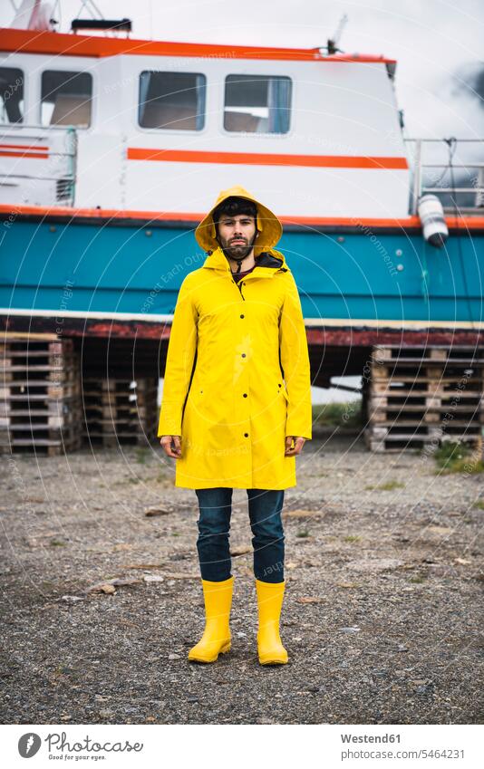 Junger Mann steht vor einem Schiff und trägt Regenkleidung, Lappland, Norwegen Regenjacke Regenjacken junger Mann junge Männer stehen stehend Gummistiefel