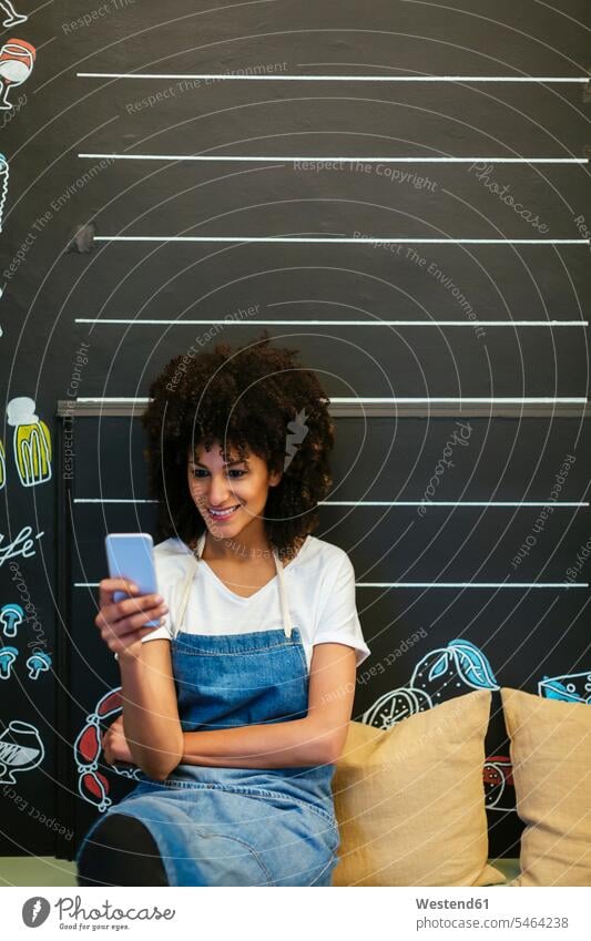 Lächelnde Frau sitzt auf einer Bank in einem Geschäft und benutzt ein Mobiltelefon Handy Handies Handys Mobiltelefone Sitzbänke Bänke Sitzbank lächeln Shop