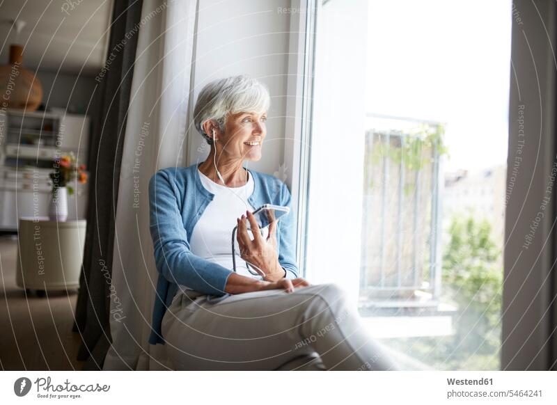 Aktive ältere Frau, die mit einem Smartphone Musik hört, während sie zu Hause sitzend aus dem Fenster schaut Farbaufnahme Farbe Farbfoto Farbphoto Innenaufnahme