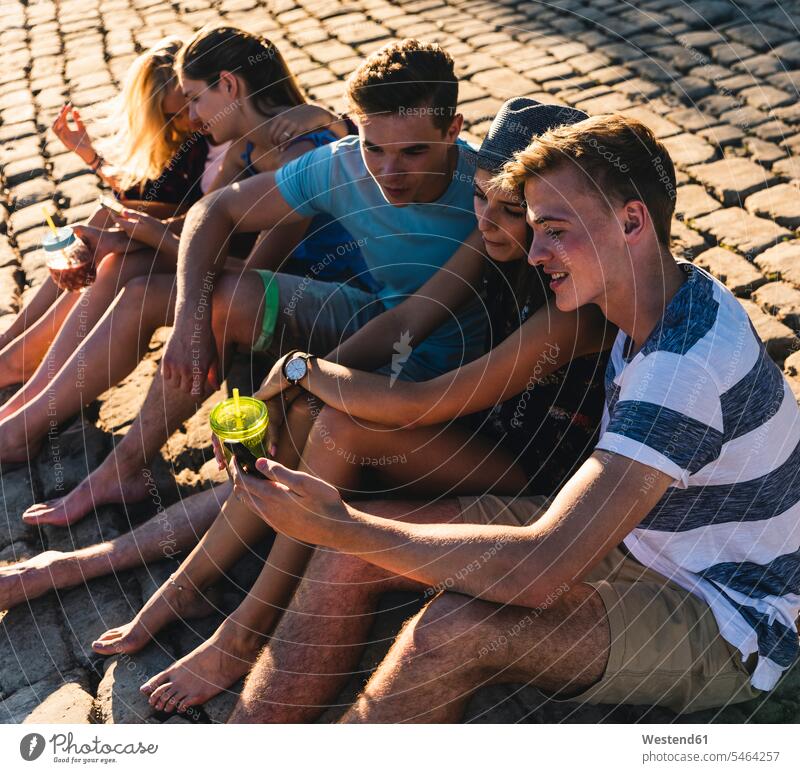 Gruppe von Freunden sitzt auf Kopfsteinpflaster mit erfrischenden Getränken und Mobiltelefonen Erfrischungsgetränk Erfrischungsgetraenk Softdrink