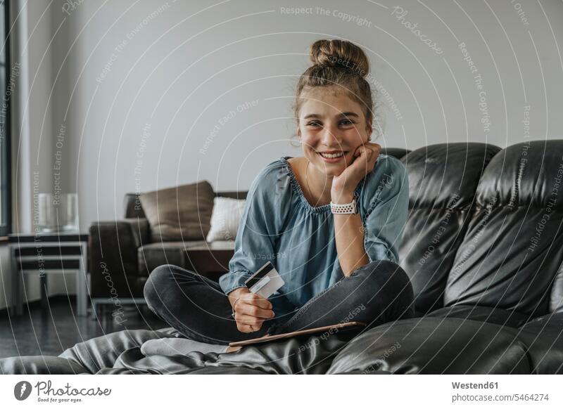 Lächelndes Mädchen mit digitalem Tablett und Kreditkarte sitzt zu Hause auf dem Sofa Farbaufnahme Farbe Farbfoto Farbphoto Deutschland Innenaufnahme