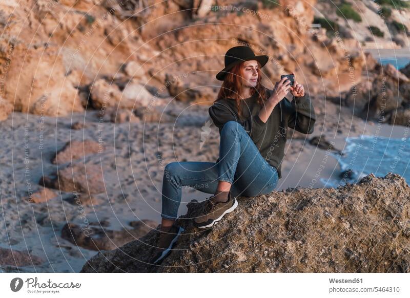 Junge Frau benutzt Smartphone am Strand bei Sonnenuntergang, Ibiza Bilder Bildnis Fotos Hüte sitzend sitzt abends freuen Glück glücklich sein glücklichsein