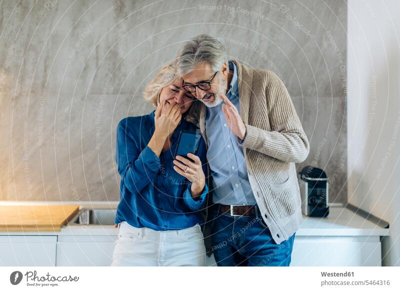 Ein erwachsenes Paar, das zu Hause in der Küche gemeinsam ein Mobiltelefon benutzt Telekommunikation telefonieren Handies Handys Mobiltelefone begruessen