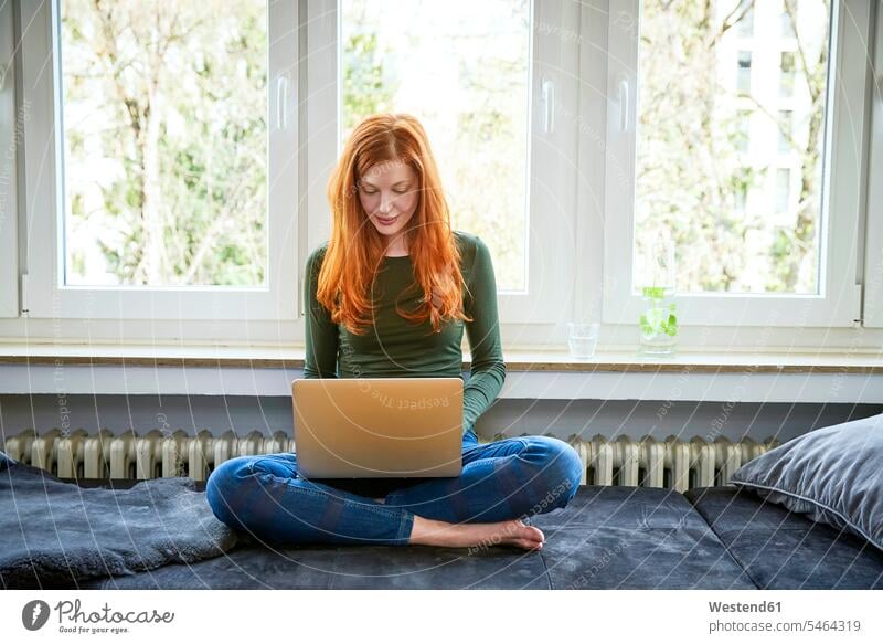 Rothaarige Frau sitzt mit Laptop vor dem Fenster weiblich Frauen sitzen sitzend benutzen benützen Notebook Laptops Notebooks Erwachsener erwachsen Mensch