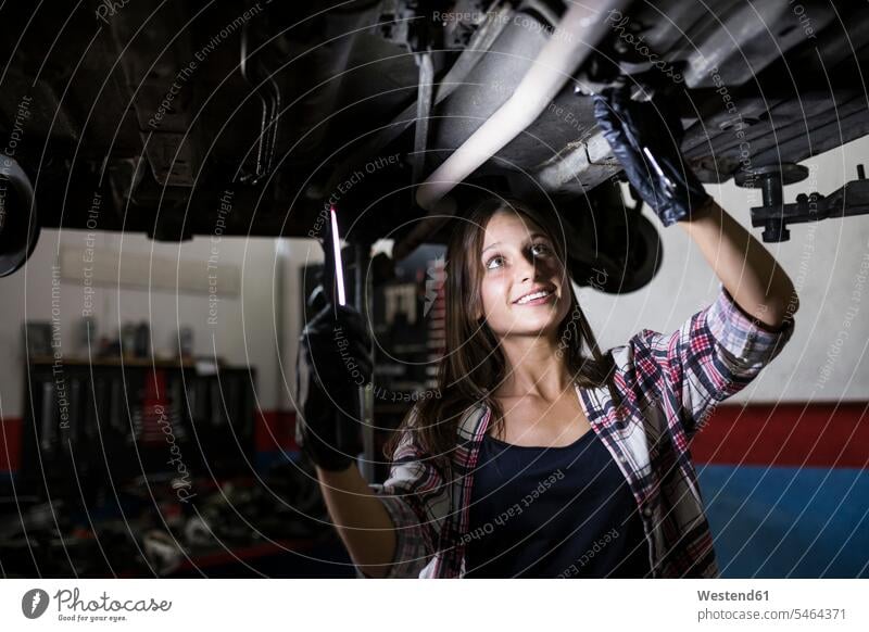 Lächelnde Frau mit Lampe, die ein Auto repariert, das unter dem Boden in einer Autoreparaturwerkstatt steht Job Berufe Berufstätigkeit Beschäftigung Jobs KFZ