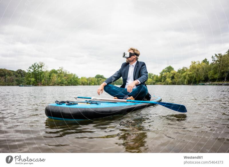 Geschäftsmann mit VR-Brille sitzt auf einem SUP-Brett auf einem See geschäftlich Geschäftsleben Geschäftswelt Geschäftsperson Geschäftspersonen Businessmann