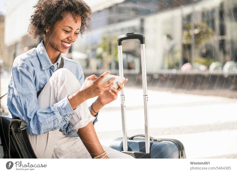 Glückliche junge Frau sitzt auf einer Bank mit Koffer und benutzt ihr Handy Leute Menschen People Person Personen 1 Ein ein Mensch nur eine Person single