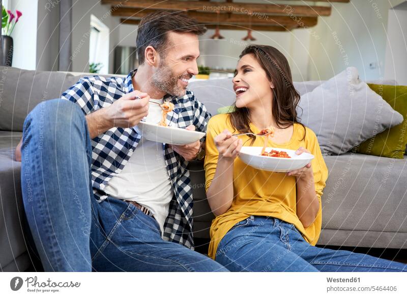 Ehepaar sitzt im Wohnzimmer und isst Spaghetti Teller Wohnraum Wohnung Wohnen Wohnräume Wohnungen essen essend lachen Spagetti sitzen sitzend Paar Pärchen Paare